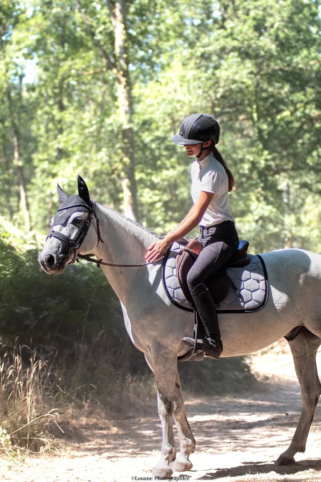 Cavalière en selle sur un cheval gris, tapis de selle gris chiné, chemin boisé