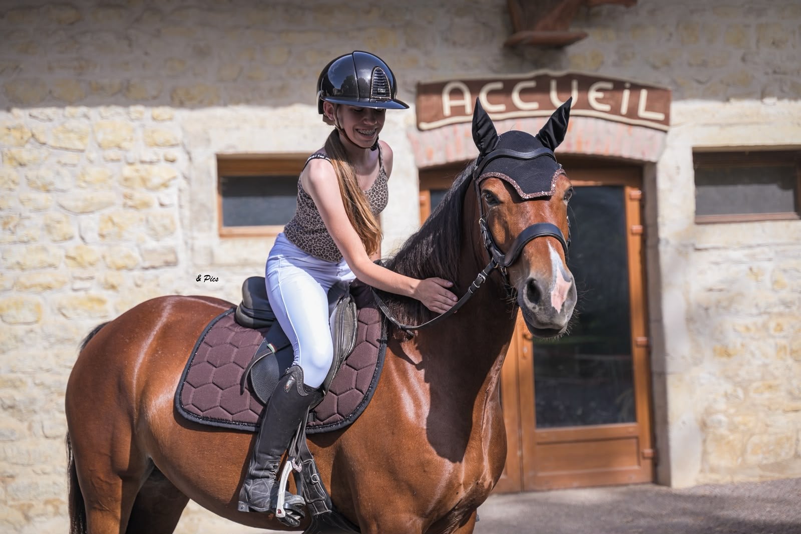 Jeune cavalière sur un cheval bai devant un bâtiment en pierre, tapis de selle chocolat