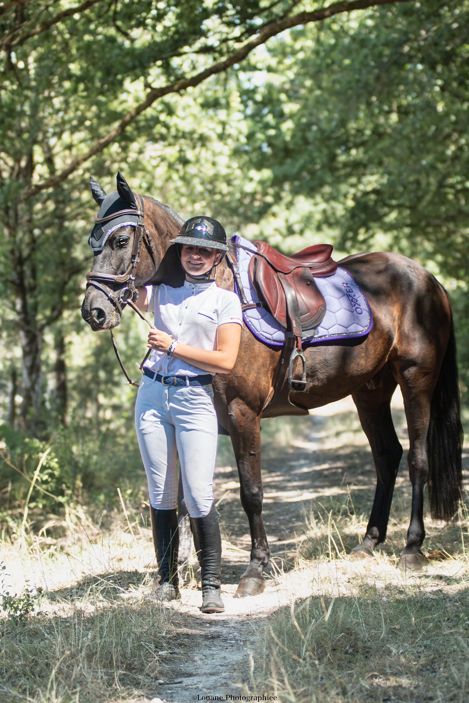 Cavalière tenant un cheval bai sur un chemin, tapis de selle lilas