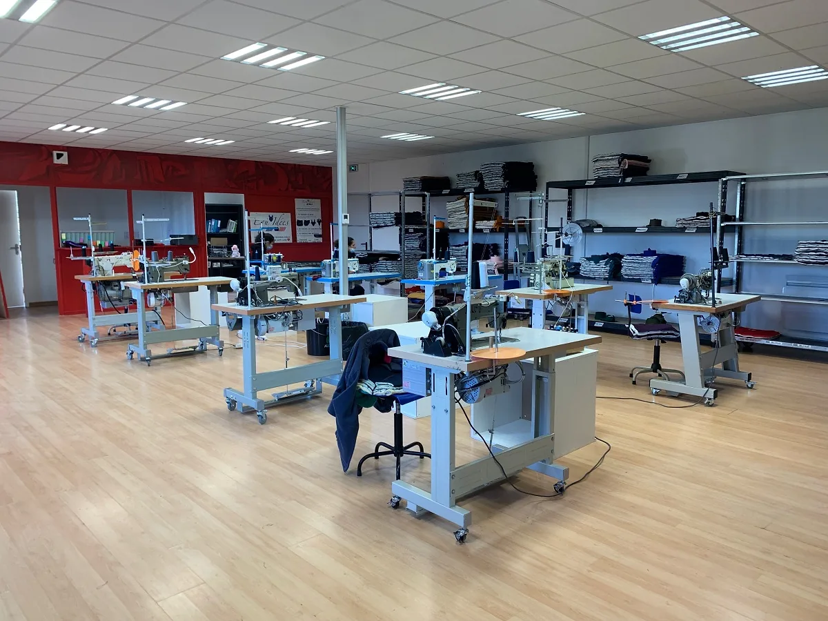 L'atelier : des machines à coudre alignées sur un parquet clair, des étagères de tissus pliés et, au fond, deux couturières devant un mur rouge au logo Equ'Idées