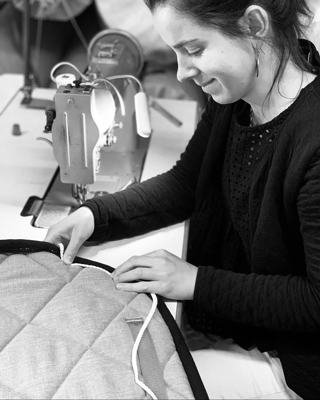 En noir et blanc, une couturière souriante guide une cordelette le long du bord d'un tapis de selle matelassé, devant sa machine à coudre